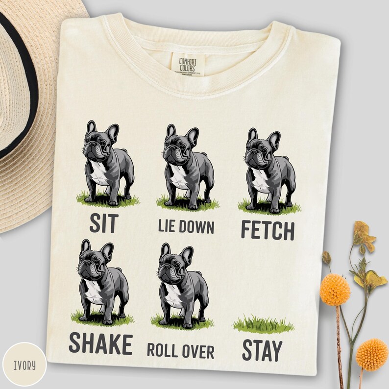 Op de afbeelding: Ivoor kleurig T-shirt met illustraties van een Franse Bulldog in verschillende poses: zitten, liggen, apporteren, schudden, rollen en blijven. De tekst onder elke hond beschrijft de actie. Het shirt heeft een Comfort Colors label.