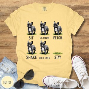Op de afbeelding: Bottergeel T-shirt met een afbeelding van zes Franse Bulldogs die commando's demonstreren: zit, ga liggen, apporteer, schud, rol om en blijf. Het shirt wordt gecombineerd met een spijkerbroek, zonnebril en witte sneakers.
