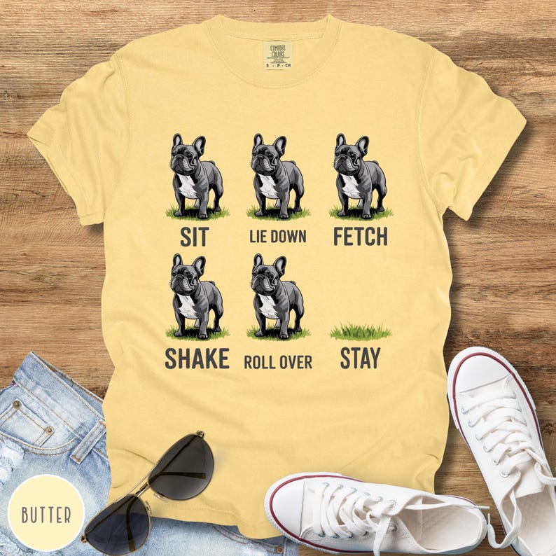 Op de afbeelding: Bottergeel T-shirt met een afbeelding van zes Franse Bulldogs die commando's demonstreren: zit, ga liggen, apporteer, schud, rol om en blijf. Het shirt wordt gecombineerd met een spijkerbroek, zonnebril en witte sneakers.