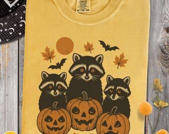 Op de afbeelding: Mosterdkleurig T-shirt met een Halloween-ontwerp. De afbeelding bevat drie wasberen, drie pompoenen, vleermuizen, een zon en herfstbladeren. Het shirt heeft de tekst "Comfort Colors" en maat informatie.