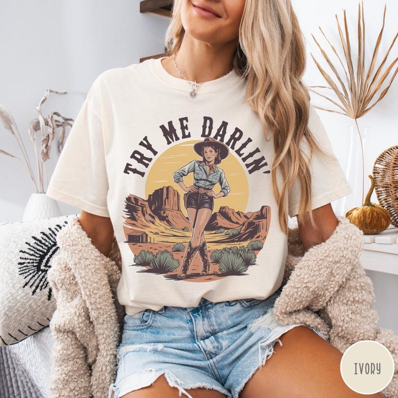 Pu&ograve; includere: T-shirt color avorio con grafica di una donna con cappello e stivali da cowboy, ambientata in un paesaggio desertico con la scritta "TRY ME DARLIN'". La maglietta ha una vestibilit&agrave; ampia.