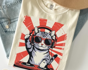 May include: Ivory-colored t-shirt featuring a cartoon cat wearing red sunglasses and headphones, with a red and white sunburst design. The cat is positioned behind a red DJ turntable. The shirt has the Comfort Colors label.