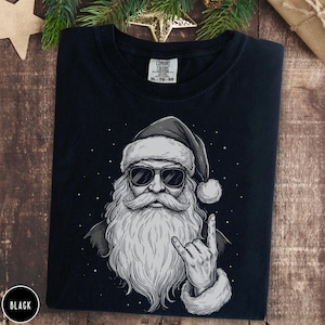 Puede incluir: Camiseta negra con un gráfico en blanco y negro de Papá Noel con gafas de sol y haciendo el gesto de la mano del rock and roll. La ilustración de Papá Noel tiene una larga barba y un sombrero. La camiseta tiene una etiqueta Comfort Colors.