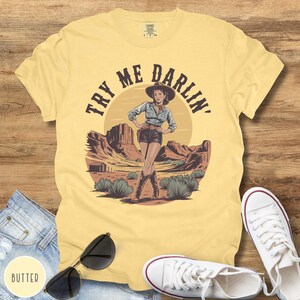 Pu&ograve; includere: T-shirt giallo burro con una grafica di una cowgirl in un paesaggio desertico con la scritta "Try Me Darlin'". Il design include una cowgirl con cappello, stivali e pantaloncini. Sono visibili anche occhiali da sole, jeans e scarpe da ginnastica bianche.