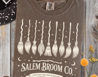 Könnte beinhalten: Espressofarbenes T-Shirt mit einer Grafik von neun stilisierten weißen Besen. Der Text "Salem Broom Co." steht darunter, mit "Crafting Enchanted Brooms Since 1692" darunter. Das Shirt ist XL-TG-EG.