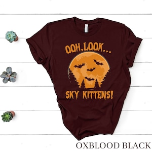 May include: A maroon t-shirt with a graphic of a full moon and bats. The text on the shirt reads "Ooh, look... Sky Kittens!"