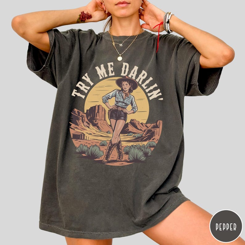 Pu&ograve; includere: T-shirt grigio scuro con un'illustrazione di un paesaggio desertico e una donna con cappello da cowboy. Il testo "TRY ME DARLIN'" &egrave; sopra l'immagine. La maglietta ha un aspetto vintage sbiadito.