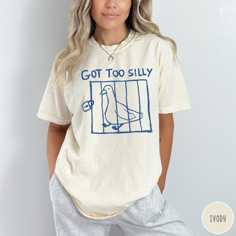 Op de afbeelding: Ivoor kleurig T-shirt met de tekst "GOT TOO SILLY" boven een blauwe illustratie van een duif in een kooi. Het shirt heeft korte mouwen en een ronde hals.