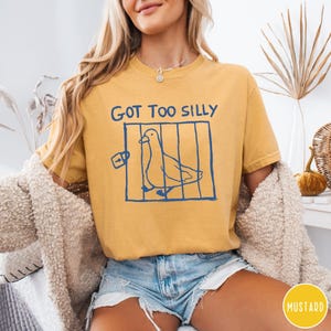 Op de afbeelding: Mosterdgeel T-shirt met de tekst "GOT TOO SILLY" en een blauwe illustratie van een vogel in een kooi. Het T-shirt is een casual stijl, geschikt voor dagelijks gebruik.