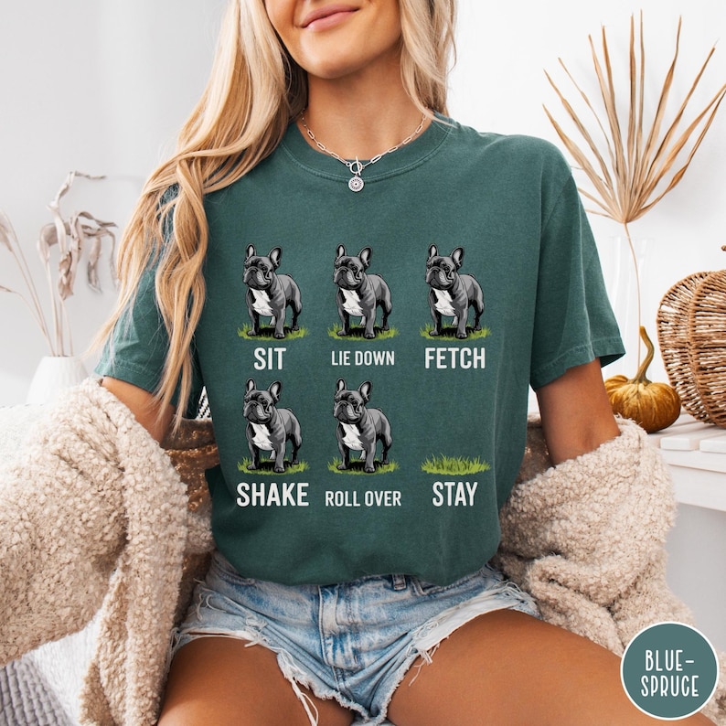 Op de afbeelding: Teal groene t-shirt met zes illustraties van een Franse Bulldog die commando's demonstreert: zit, ga liggen, apporteer, schud, rol om en blijf. Elke hondenillustratie is in zwart-wit, met het commando eronder geschreven.
