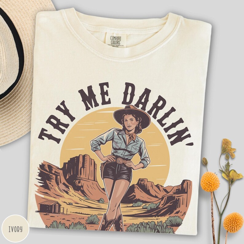 Pu&ograve; includere: T-shirt color avorio con grafica di una donna con cappello da cowboy e la scritta "TRY ME DARLIN'". Il design presenta un paesaggio desertico con montagne e un sole al tramonto. La maglietta &egrave; piegata e ha un'etichetta Comfort Colors.