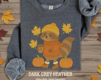 Pode incluir: Camisola unissexo cinza escuro mesclado com um guaxinim de desenho animado usando um chapéu amarelo e um suéter laranja, segurando uma xícara de café e em pé ao lado de duas abóboras. Folhas de bordo amarelas circundam o guaxinim. O texto "DARK GREY HEATHER" está na parte inferior.