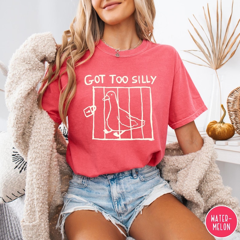 Op de afbeelding: Een koraalkleurig T-shirt met de tekst "GOT TOO SILLY" boven een witte afbeelding van een vogel in een kooi. Het shirt wordt gecombineerd met een spijkerbroek. Het woord "WATERMELON" staat in een roze cirkel rechtsonder.