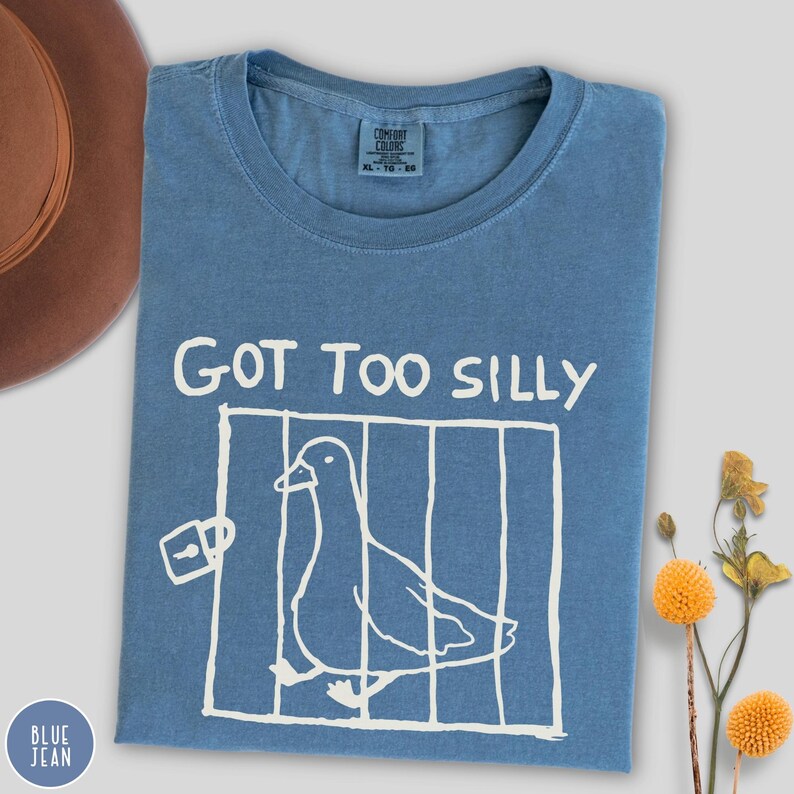 Op de afbeelding: Blauw spijkerbroekkleurig T-shirt met de witte tekst "GOT TOO SILLY". Een witte afbeelding van een vogel in een kooi staat onder de tekst. Het shirt heeft een ronde hals en korte mouwen. Het Comfort Colors-label is zichtbaar.