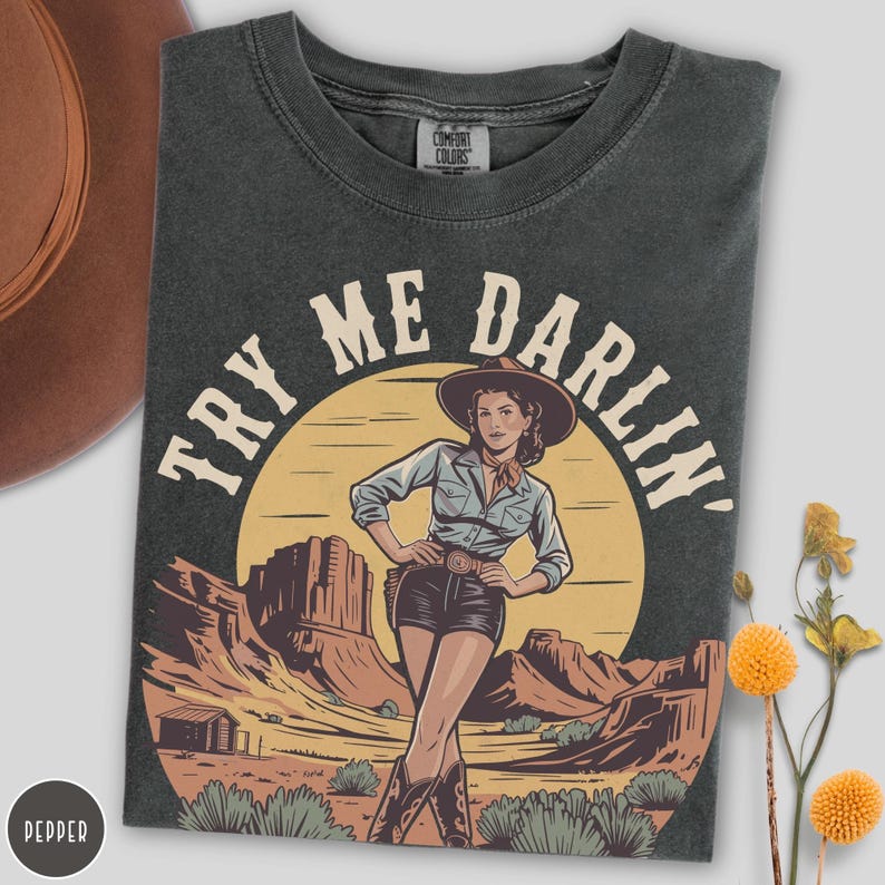 Pu&ograve; includere: T-shirt grigio scuro con grafica di una cowgirl davanti a un paesaggio desertico e la scritta "TRY ME DARLIN'". La cowgirl indossa un cappello, una camicia blu e pantaloncini neri. La maglietta &egrave; del marchio Comfort Colors.