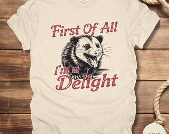 May include: Ivory-colored t-shirt featuring a black and white illustration of an opossum with its tongue out. The text "First Of All I'm a Delight" is printed in a burgundy color above and below the opossum graphic. The shirt is made of a soft material.