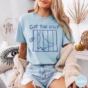Op de afbeelding: Lichtblauw T-shirt met de tekst "GOT TOO SILLY" boven een blauwe illustratie van een vogel in een kooi. Het shirt heeft een casual stijl.