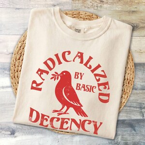 May include: Cream-coloured t-shirt featuring a red dove graphic holding an olive branch, with the text "RADICALIZED BY BASIC DECENCY". The shirt is folded on a woven mat.