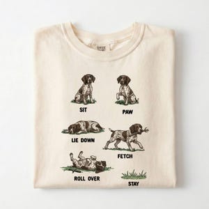 May include: Cream-colored t-shirt featuring illustrations of a brown and white dog demonstrating commands: sit, paw, lie down, fetch, roll over, and stay. The shirt has a crew neck and the brand name "Comfort Colors" is visible.