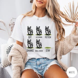 Op de afbeelding: Wit T-shirt met zes illustraties van een Franse Bulldog die commando's uitvoert: zit, ga liggen, apporteer, schud, rol om en blijf. De tekst onder elke hond beschrijft de actie. Een casual kledingstuk voor dagelijks gebruik.
