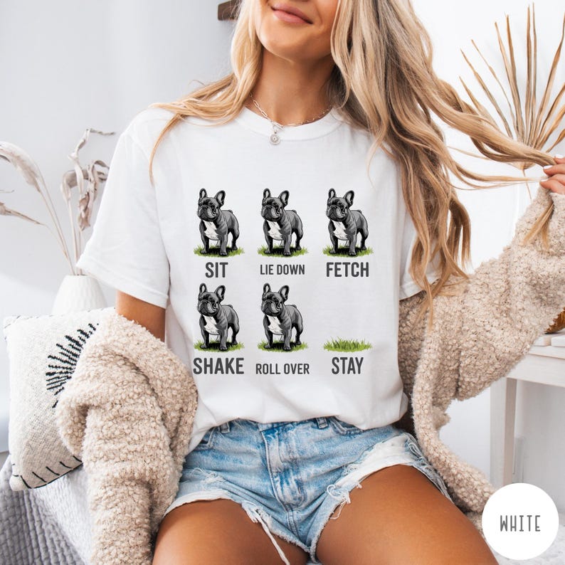 Op de afbeelding: Wit T-shirt met zes illustraties van een Franse Bulldog die commando's uitvoert: zit, ga liggen, apporteer, schud, rol om en blijf. De tekst onder elke hond beschrijft de actie. Een casual kledingstuk voor dagelijks gebruik.