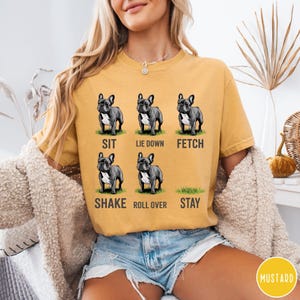 Op de afbeelding: Mosterdkleurig T-shirt met zes illustraties van een Franse Bulldog die commando's uitvoert: zit, ga liggen, apporteer, schud, rol om en blijf. De tekst voor elk commando staat onder elke hondenillustratie.