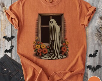 May include: Orange t-shirt featuring a graphic of a ghost in a window with flowers. The ghost is draped in a flowing sheet, and the window is framed by potted flowers. The shirt is a solid orange color, with rolled-up sleeves.