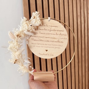 Peut inclure: Un cadre en bois avec un anneau doré et des fleurs séchées. La plaque porte un texte gravé et est soutenue par un socle en bois. Le texte se lit "Eu es la nounou...Raphaël & Leo."