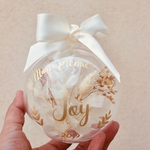 Peut inclure: Une boule de Noël en verre transparente avec un nœud en ruban blanc, des fleurs séchées et une inscription dorée qui dit "Mon premier Noël Joy 2019".