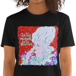 May include: Black t-shirt featuring a colorful illustration of a woman with a powdered wig and the text "Qu'ils mangent de la Brioche." The artwork is set against a red background with floral accents, creating a vintage aesthetic.