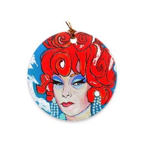 May include: Round ceramic ornament featuring a colorful illustration of a person with red curly hair, blue eyeshadow, and a blue background.