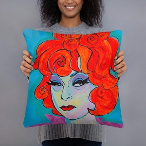 May include: A colorful throw pillow featuring a stylized portrait of a woman with bright red hair. The woman's face is painted in a vibrant palette of blues, greens, and yellows. The background is a solid turquoise blue.