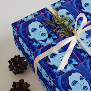 May include: Blue gift wrap featuring a repeating pattern of a woman's portrait in shades of blue. The wrapped gift is tied with a cream-colored ribbon and adorned with a sprig of greenery. Two pinecones are placed near the gift.