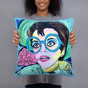 May include: A blue and green throw pillow with a colorful illustration of a woman wearing large blue glasses and a pink speech bubble that says "Sid Luft is an animal".