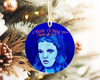 May include: A round blue Christmas ornament with a painting of Dorothy from the Wizard of Oz. The ornament has the text "I think I'll miss you most of all" in red.