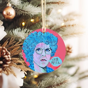May include: Round Christmas ornament with a pink background and a cartoon drawing of a woman with blue hair and glasses. The woman is saying "Well Hell" in a speech bubble.