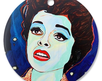 May include: A round, blue and white painted ornament featuring a portrait of a woman with dark hair and red lips. The background is a dark blue with white stars.