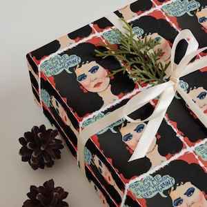 May include: Gift-wrapped present with patterned paper featuring a woman's portrait with the text "Are you calling me a Miss Valdosta Feed and Grain?". The gift is tied with a white ribbon and decorated with a sprig of greenery and two pinecones.