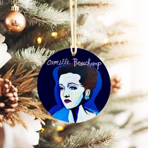 May include: A round Christmas ornament featuring a stylized portrait of Camille Beauchamp on a dark blue background. The ornament has a gold string for hanging and is displayed on a decorated Christmas tree.