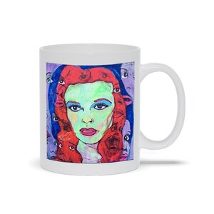 May include: White ceramic mug with a colorful illustration of a woman's face surrounded by other faces. The woman has red hair, green eyes, and a light blue background.