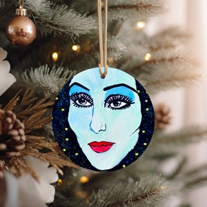 May include: A round Christmas ornament featuring a hand-painted portrait of a woman with blue eyeshadow and red lipstick. The background is a dark blue with yellow stars.