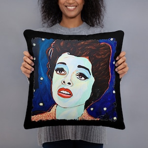 May include: A black throw pillow with a colorful painting of a woman's face. The woman has blue eyes, red lips, and dark hair. The background is a dark blue with white stars.