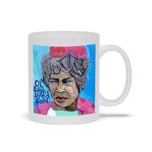 May include: White ceramic mug with a colorful illustration of a woman with gray hair wearing a pink hat. The text "Ol' fish eyed fool" is written in blue on the left side of the mug.