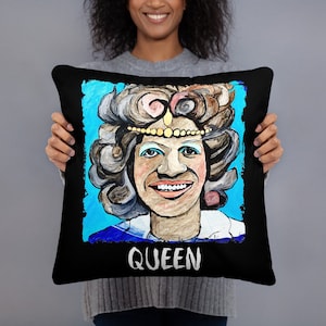 May include: Black square pillow featuring a colorful portrait of a smiling person with a crown and elaborate hairstyle. The word "QUEEN" is printed in white at the bottom of the pillow. The artwork has a painted look with a blue background.