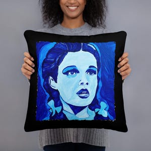 May include: Black throw pillow featuring a blue portrait of a woman with a surprised expression. The artwork is in shades of blue, with a black border. The pillow is a decorative item.