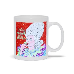 May include: White ceramic mug with a colorful illustration of a woman with a large white wig, wearing a colorful patterned dress and eating a green pastry. The text "Qu'ils mangent de la brioche" is written in green and black on a red background.