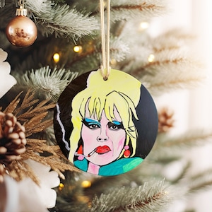 May include: Round Christmas ornament featuring a hand-painted portrait of a woman with blonde hair, wearing red lipstick and earrings. The background is black with a white smoke effect.