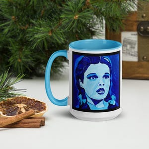 May include: A white ceramic mug with a light blue handle and interior. The mug features a blue and white portrait of a woman. The mug is sitting on a white surface with a dried orange slice and cinnamon sticks.