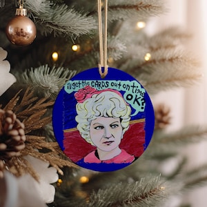 May include: A round blue Christmas ornament with a cartoon image of a woman with blonde hair and a pink shirt. The woman is saying "I'll get the cards out on time OK?" in a speech bubble.