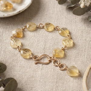 May include: A bracelet featuring translucent, pale yellow, heart-shaped gemstones linked together with gold-toned metal rings. The piece includes a gold-toned hook clasp and an extender chain that terminates in a single heart-shaped gemstone charm.